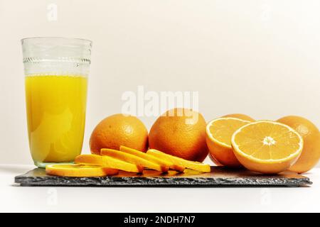 Un mazzo di arance mature con fettine e arance spaccate accanto ad un bicchiere con succo su una superficie nera Foto Stock