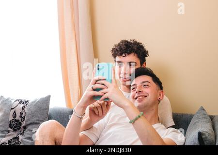 Primo piano di una coppia gay sdraiata sul divano e che utilizza il cellulare per interagire sui social media. Routine quotidiana Foto Stock