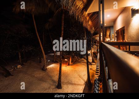 Vista dall'alto in una posizione bassa e lunga dal balcone di un hotel con tetto in paglia all'entrata della proprietà con sabbia bianca, alte palme, e. Foto Stock