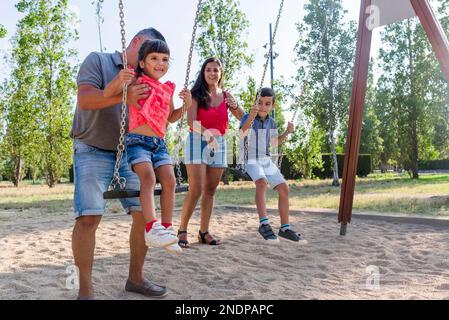 Famiglia con figlio e figlia divertirsi giocando con l'altalena in un parco. Foto Stock