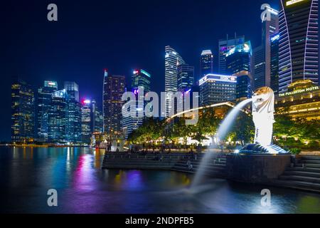 Il Merlion, simbolo nazionale di Singapore, visto di notte a Marina Bay con il Fullerton Hotel e il quartiere finanziario di Singapore sullo sfondo. Foto Stock