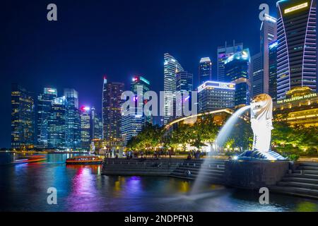 Il Merlion, simbolo nazionale di Singapore, visto di notte a Marina Bay con il Fullerton Hotel e il quartiere finanziario di Singapore sullo sfondo. Foto Stock