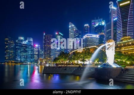 Il Merlion, simbolo nazionale di Singapore, visto di notte a Marina Bay con il Fullerton Hotel e il quartiere finanziario di Singapore sullo sfondo. Foto Stock