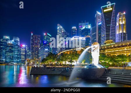 Il Merlion, simbolo nazionale di Singapore, visto di notte a Marina Bay con il Fullerton Hotel e il quartiere finanziario di Singapore sullo sfondo. Foto Stock