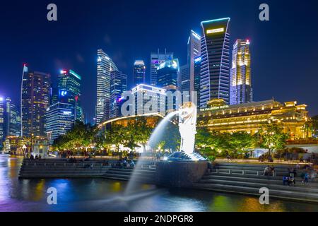 Il Merlion, simbolo nazionale di Singapore, visto di notte a Marina Bay con il Fullerton Hotel e il quartiere finanziario di Singapore sullo sfondo. Foto Stock