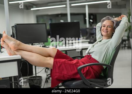 Donna caucasica matura tolse le scarpe e mise i piedi sul desktop in ufficio. Foto Stock