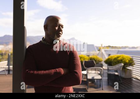 Uomo anziano afro-americano che guarda lontano mentre si trova in piedi sulla terrazza a casa. stile di vita e concetto di vita senior in pensione Foto Stock