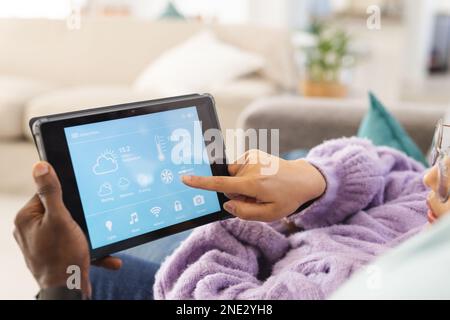 Felice coppia diversa seduto sul divano nel soggiorno e utilizzando un tablet con interfaccia domestica intelligente. Trascorrere il tempo di qualità a casa, la vita domestica e la vita Foto Stock