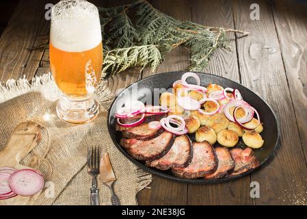 pranzo a base di prosciutto fritto con patate e cipolle in una padella accanto a un bicchiere di birra su un tavolo rustico in legno Foto Stock