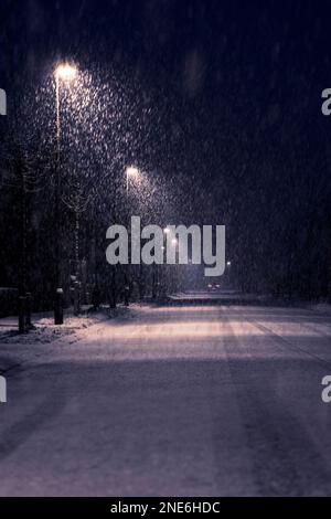 Una strada illuminata da lampioni di strada circondata da fiocchi di neve in caduta sul lato della strada. Le luci della strada illuminano la nevicata pesante di notte e. Foto Stock