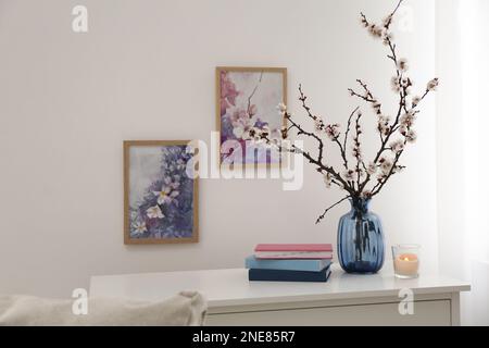 Albero fiorito ramoscelli in vaso di vetro, candela bruciante e libri sul tavolo vicino parete bianca a casa Foto Stock