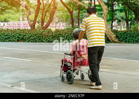 Vista posteriore dell'uomo anziano asiatico che cammina con la donna anziana disabile seduta in sedia a rotelle all'aperto indossando maschere mediche. L'uomo spinge la vecchia signora in carriola Foto Stock