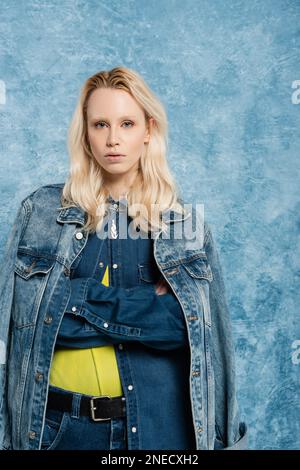 donna bionda in giacca denim posa con braccia incrociate vicino blu textured sfondo, immagine stock Foto Stock