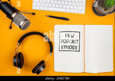 Vista dall'alto del testo NUOVO EPISODIO PODCAST su blocco note su scrivania con microfono, tastiera per computer e cuffie, concetto di podcasting Foto Stock