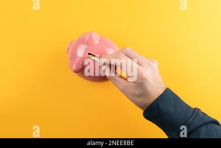 risparmio di denaro e il concetto di profitto, vista dall'alto della mano inserendo moneta dorata in banca rosa piggy contro sfondo arancione colorato Foto Stock