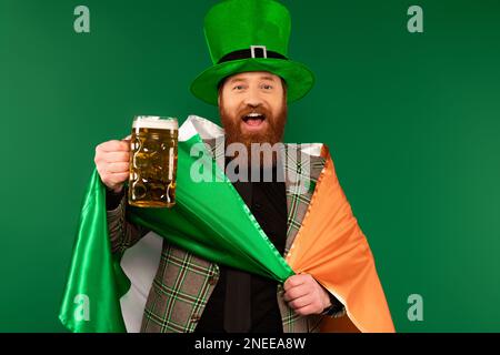 Allegro uomo portatore di birra e bandiera irlandese durante il giorno di san patrizio isolato su verde, immagine stock Foto Stock