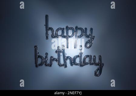 Scritta in argento con palloncino di buon compleanno e luce blu creativa. Foglio di lettere su sfondo orizzontale. Saluto metallico, decorazioni per feste, inviti a eventi. Foto Stock