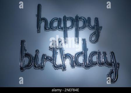 Scritta in argento con palloncino di buon compleanno e luce blu artistica. Foglio di lettere su sfondo orizzontale. Saluto metallico, decorazioni per feste, inviti a eventi. Foto Stock