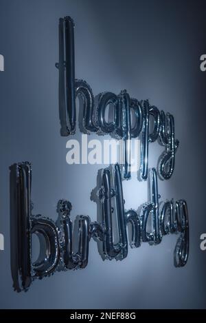 Scritta in argento con palloncino di buon compleanno e luce blu. Foglio di lettere da lato su sfondo verticale. Saluto metallico, decorazioni per feste, inviti a eventi. Foto Stock