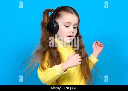 Felice bambina con cuffie wireless su sfondo blu che fa la danza si muove verso la musica in cuffie, bambino con capelli lunghi. Foto Stock