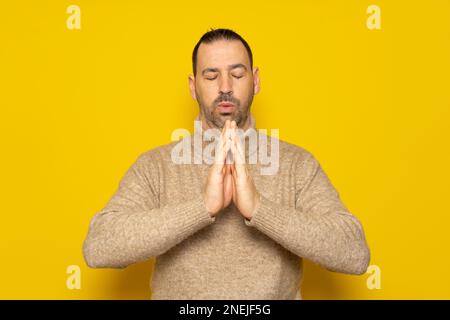 Barbuto uomo ispanico indossando dolcevita beige isolato su sfondo giallo, tenendo le mani in preghiera o meditazione, guardando rilassato e tranquillo Foto Stock