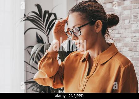 Mal di testa. Foto ravvicinata di una giovane donna, seduta su un divano con gli occhi chiusi, che tocca la testa mentre soffre di emicrania. Business Foto Stock