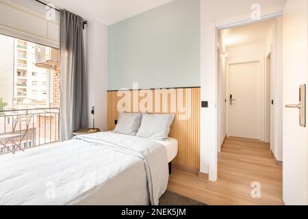 Letto matrimoniale in camera, accanto a una grande finestra a parete con accesso a un balcone e vista sulle case vicine. Ci sono delle tende sulla finestra Foto Stock