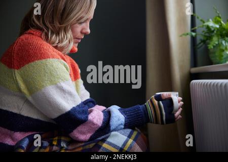 Donna in guanti con bevanda calda cercando di mantenere caldo dal radiatore durante il costo di Living Energy crisi Foto Stock