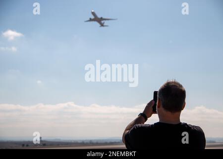 Un uomo adulto un po' calvo vestito di nero che guarda e punta mentre gli aerei decolgono dalla pista dell'aeroporto Foto Stock