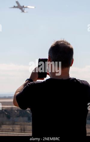 Un uomo adulto un po' calvo vestito di nero che guarda e punta mentre gli aerei decolgono dalla pista dell'aeroporto Foto Stock