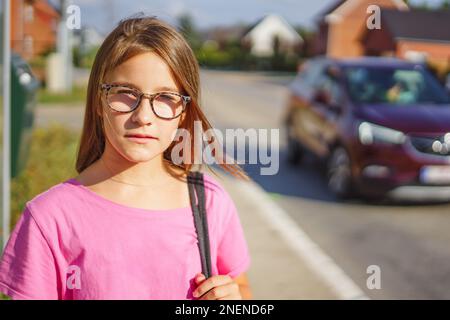 Ritratto di una ragazza carina in occhiali per la visione, guarda nella fotocamera, tiene una borsa sulla spalla e aspettare vicino alla strada in estate giorno di sole. Foto Stock