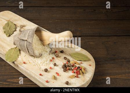 Riso in un sacchetto di iuta con un cucchiaio di legno sul tavolo da cucina. Nelle vicinanze su una foglia di alloro del tagliere e diversi tipi di pepe Foto Stock