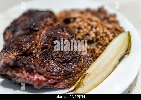 Bistecca di ribeye cotta macro closeup con taglio spesso e crosta marrone su piatto bianco con sottaceti di aneto e grano saraceno sullo sfondo Foto Stock