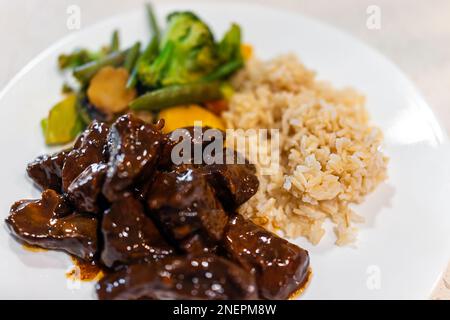 Pezzi di carne cotti di cuore di manzo cosparsi in semi di sesamo e salsa con riso marrone e broccoli mescolare-friggere verdure macro closeup su cena bianca lui Foto Stock