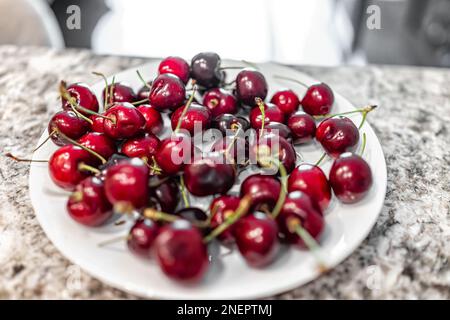Grandi ciliegie dolci fresche lavate su piatto bianco di colore rosso intenso e gambi verdi in primo piano con nessuno sul tavolo isola cucina Foto Stock