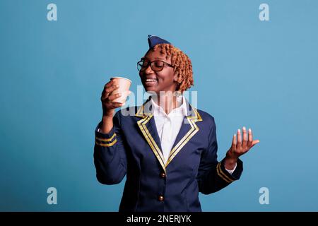 Studio ritratto di professionista afro-americana femminile di volo attendente in uniforme con tazza di caffè caldo usa e getta. Abiti formali in piedi sorridenti su sfondo blu. Foto Stock