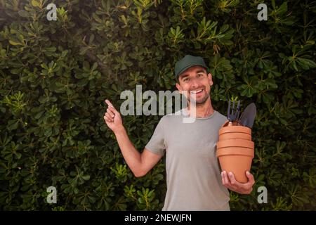 L'uomo giardiniere in tappo verde tiene pentole di terracotta con forniture di giardinaggio in mano. Punta il dito verso la recinzione sempreverde della matifolia di phillyrea. H Foto Stock