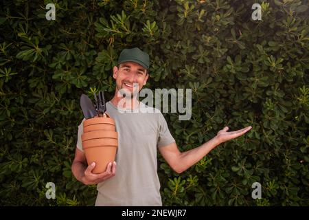 L'uomo giardiniere in tappo verde tiene pentole di terracotta con forniture di giardinaggio in mano. Punta il dito verso la recinzione sempreverde della matifolia di phillyrea. H Foto Stock