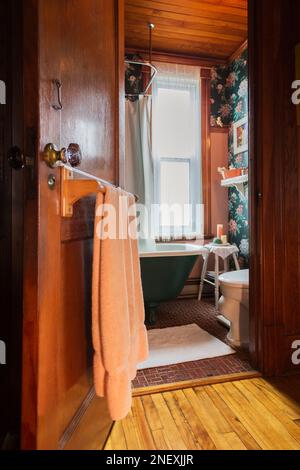 Porta aperta del bagno principale con vasca verde e bianca, pavimento in piastrelle di ceramica rossa e soffitto e pareti in legno di pino colombiano nella vecchia casa. Foto Stock