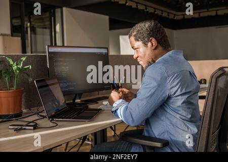 Black Man scrive righe di codice su PC desktop con due monitor e un notebook a parte in elegante ufficio. Programmazione professionale per sviluppatori maschili Foto Stock