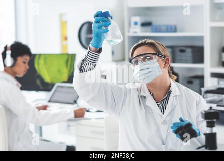 HMM, i colori cambiarono... una giovane e attraente scienziata che analizzava un liquido in un laboratorio con la sua collega sullo sfondo. Foto Stock