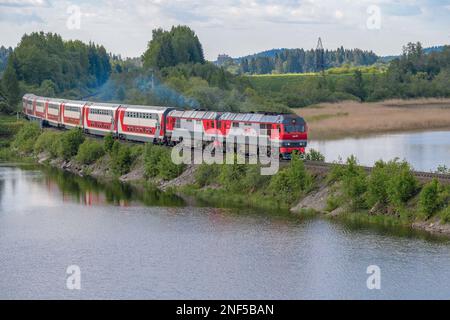 SORTAVALA, RUSSIA - 11 GIUGNO 2022: Un moderno treno passeggeri a due piani con due locomotive diesel TEP-70BS si avvicina alla diga del lago di Karmalanyarvi o Foto Stock