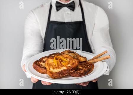 Uno chef cameriere in grembiule e cravatta che mostra un piatto di orrija gourmet. Un dolce tipico spagnolo fatto con pane, latte, uova, zucchero e cannella che io Foto Stock