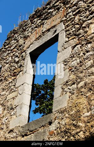 Un muro di pietra dall'aspetto medievale che mostra una finestra attraverso la quale si possono vedere rami di un albero e un cielo blu indaco senza nuvole. Foto Stock