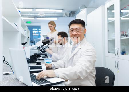 Ritratto di tecnico asiatico di laboratorio, uomo con gruppo di persone che lavorano in laboratorio di ricerca, uomo con microscopio in camice bianco e occhiali di protezione sorridendo e guardando la macchina fotografica. Foto Stock
