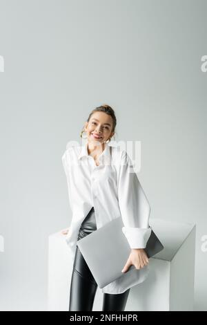 donna gioiosa in orecchini a cerchio e camicia oversize che tiene il laptop e guardare la macchina fotografica vicino al cubo bianco su sfondo grigio, immagine stock Foto Stock