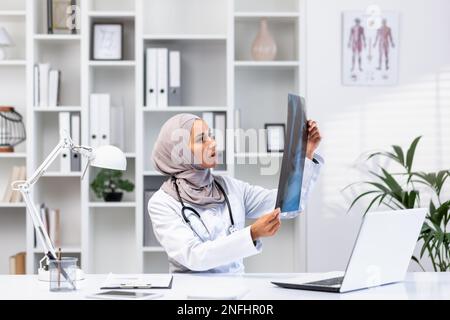 Giovane medico femminile di successo in hijab che lavora in clinica all'interno di un ufficio medico, donna musulmana è arrabbiata e pensare immagine a raggi X seduto sul posto di lavoro con il computer portatile. Foto Stock