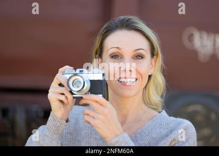 Vista frontale di una donna sorridente di mezza età che tiene in mano una vecchia macchina fotografica indossando un maglione marrone di fronte a un vecchio vagone ferroviario Foto Stock