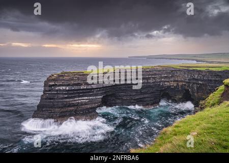 Solitario pescatore in piedi sul bordo delle scogliere di Downpatrick Head con onde che si schiantano sotto all'alba. Spettacolare alba con cielo moso, Mayo, Irlanda Foto Stock