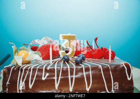 Torta di compleanno al cioccolato con bacche e numero tre candele d'oro su sfondo blu, copia space.greetings,A candela numero è acceso in celebrazione di Foto Stock